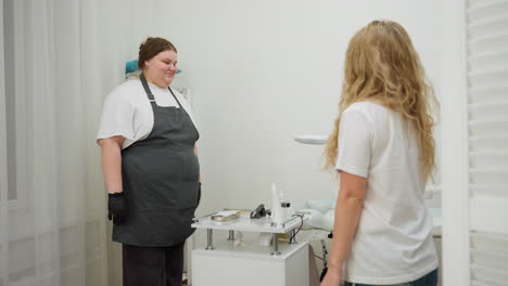 beautician in apron welcomes client with friendly smile while standing near treatment bed and tool station in clean, softly lit salon environment with sheer curtains and white decoration