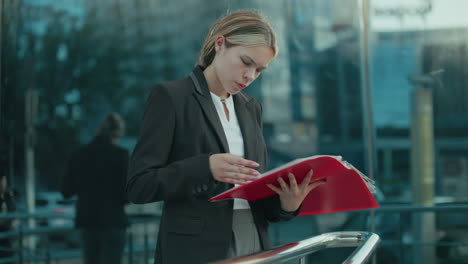 female ceo reviewing red folder near iron railing with glass office building reflecting city surroundings and sunlight ambiance, portraying modern leadership