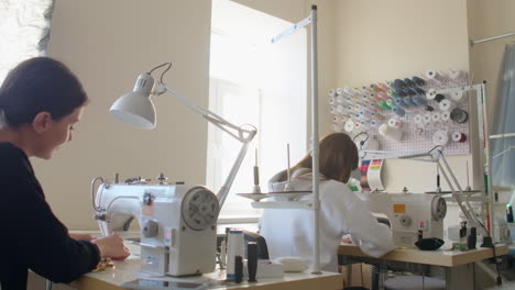 Two-women-seamstresses-working-on-sewing-machines-in-the-tailoring-workshop-of-the-garment.-The-creation-of-designer-dresses.-Small-business.-Two-employees-work-on-sewing-machines-from-the-back