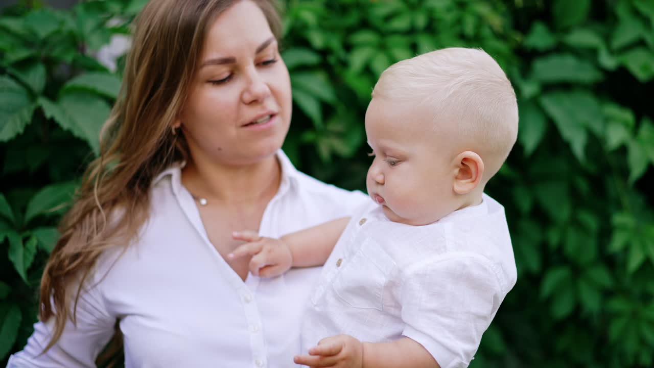 Long-haired Caucasian woman holding a blond baby boy outdoors. Mom talks to her child giving him a yellow flower.