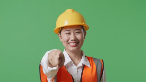 close up of asian female engineer with safety helmet smiling and touching her chest then pointing at you while standing in the green screen background studio