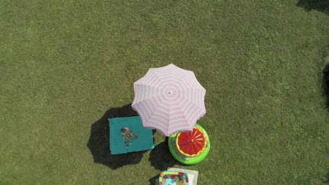 top-down shot of an adorable toddler sitting on the light green picnic mat under the beautiful pink umbrella in the lush garden - ascending drone