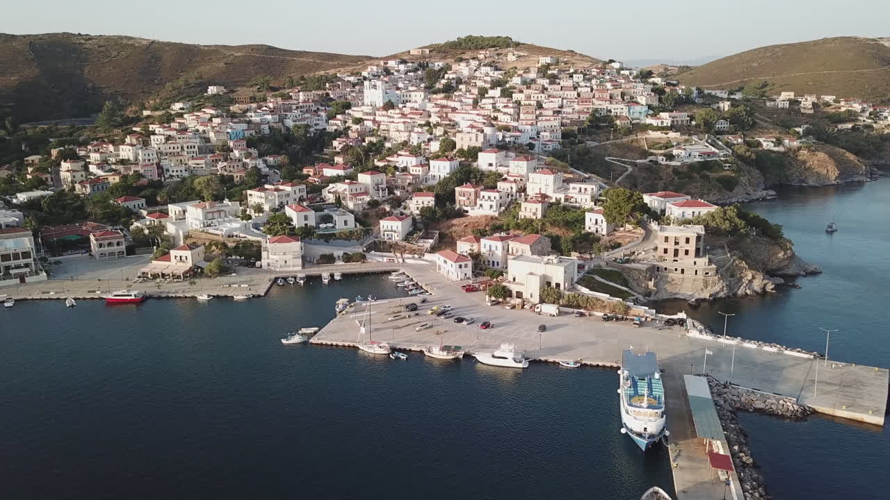 Greece, Ionusses Island, Drone Footage of the beautiful harbor, town with white houses and the landscape on a calm day. A vessel is abreast to the dock and couple of sailboats and small boats.