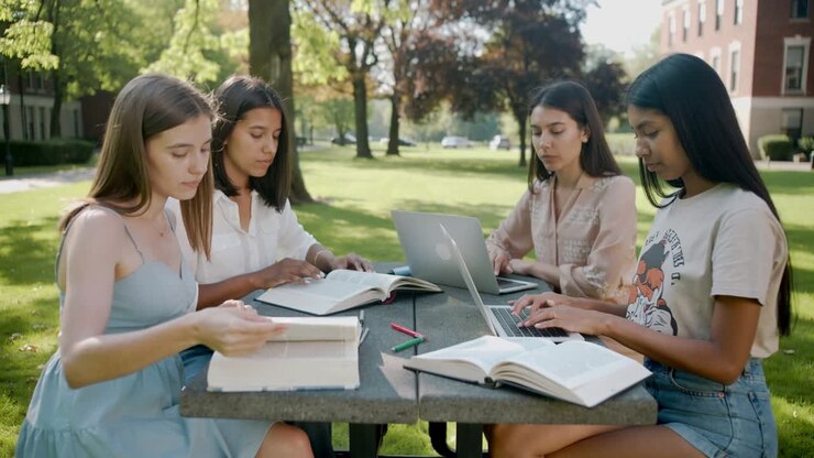 Quattro studenti in abiti casual studiano sul prato universitario, sfogliando pagine e digitando con libri di testo e un laptop.