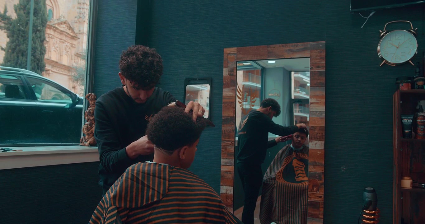 Beautiful slow motion shot of a wide shot of a haircut by a professional barber in a barber shop styling a young afro man with scissors during the afternoon