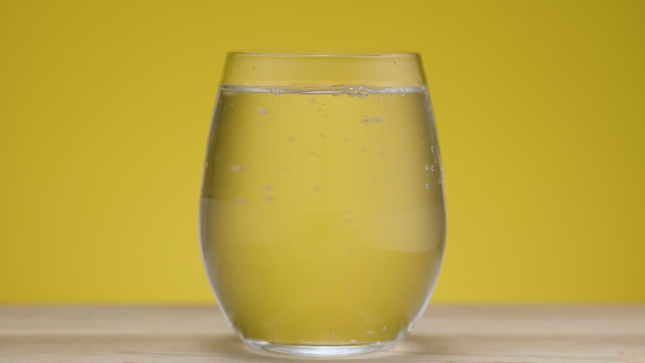 Inquadratura statica di un bicchiere d'acqua trasparente con acqua frizzante, isolato su sfondo giallo.