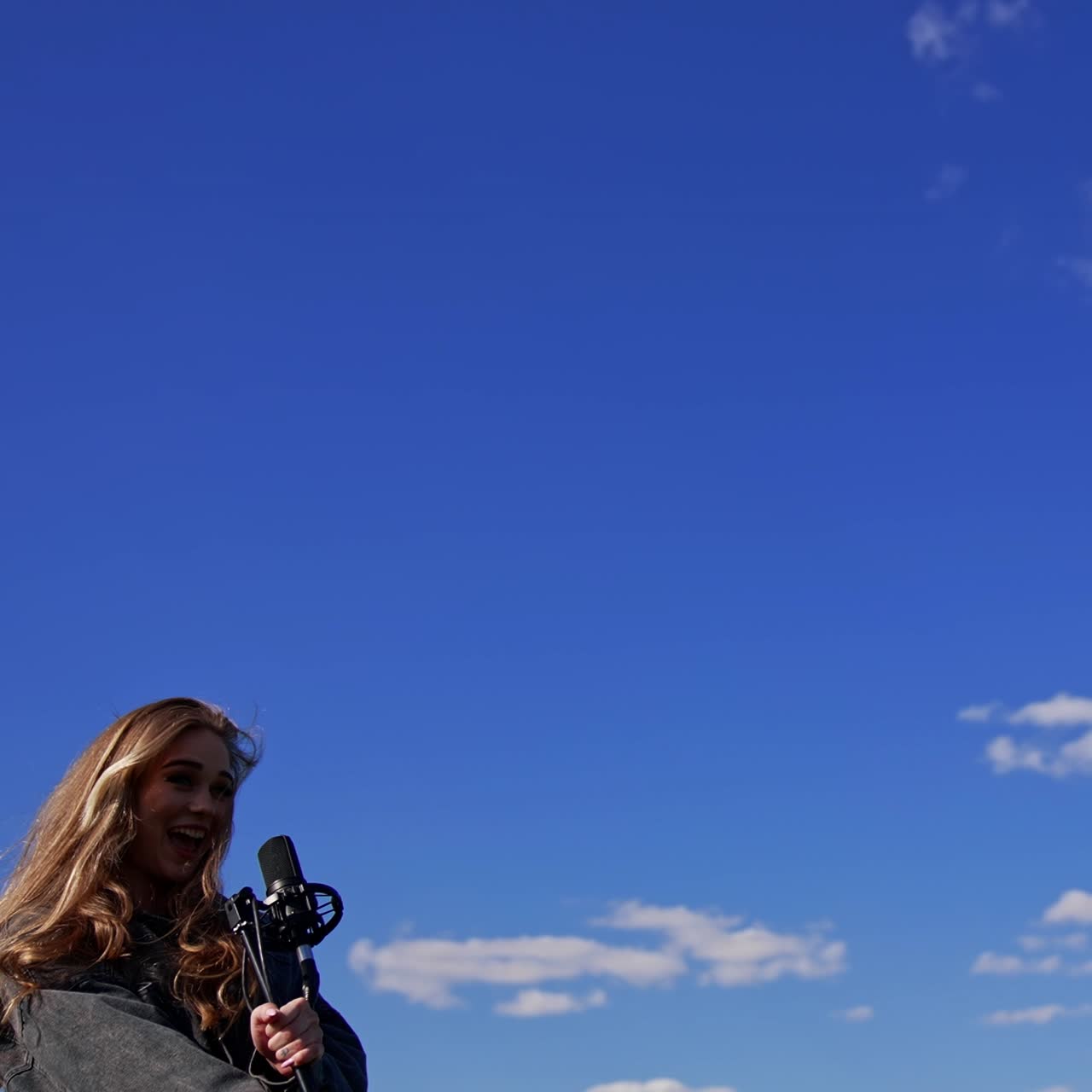 Beautiful female performing a song alone outdoors. Young singer girl with a microphone on blue sky background. View from below