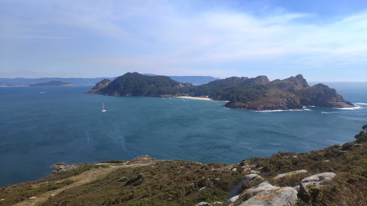 Mountainous island with beach and sailboat sailing in front on a sunny day, panoramic shot roll to the right, Cíes island, Pontevedra, Galicia, Spain