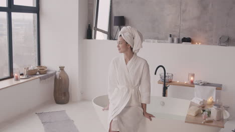 front view of a woman sitting on the edge of bathtub posing and smiling at the camera with a towel on her head and a bathrobe