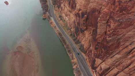 aerial view of black vehicle on deserted road by colorado river under high steep cliff in utah landscape, tilt down drone shot