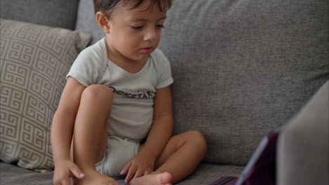 Nice-latin-toddler-in-a-light-blue-baby-romper-sitting-on-a-grey-couch-watching-cartoons-on-a-purple-tablet