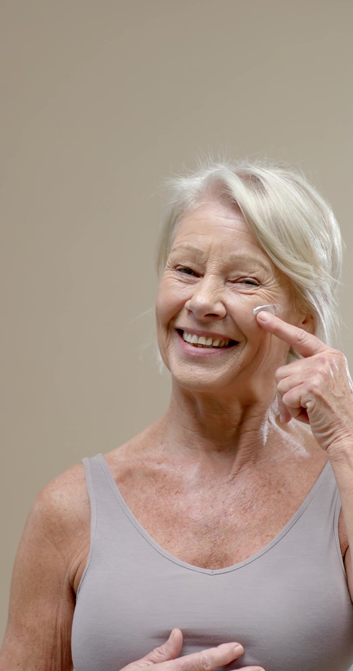 Video verticale: Donna anziana che tiene la crema sulla punta delle dita, applicandola sulla guancia sinistra in studio per la cura della pelle
