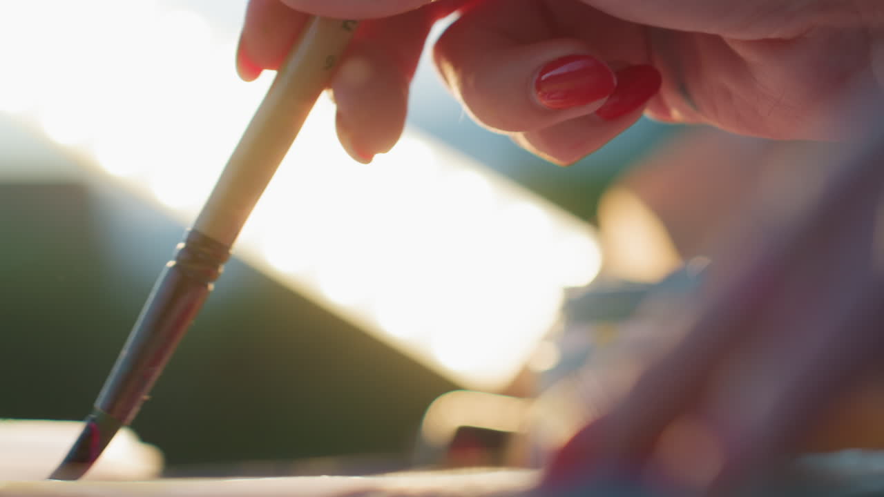 close up view of craft woman hand with red painted nails holding brush dipping into paint and applying colorful strokes on paper with soft blurred background and evening light texture details