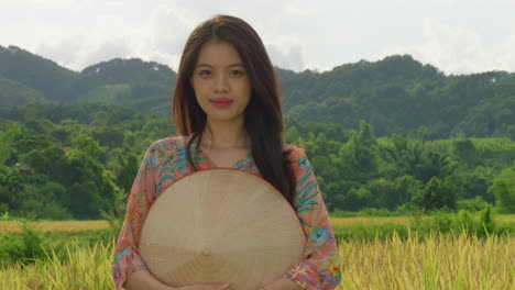 Joven-Agricultora-Vietnamita-Parada-Frente-A-Una-Plantación-De-Arroz-Con-Ropa-Tradicional-Y-Sombrero-De-Bambú-Sonriendo-A-La-Cámara-En-Cámara-Lenta