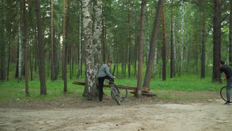 back view of cyclist riding on an untarred road surrounded by tall birch trees in a forest, the cyclist is wearing a grey jacket, parked close to a tree, with another rider following from behind