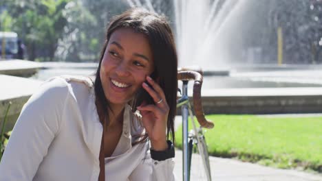mujer afroamericana con bicicleta hablando en un teléfono inteligente sentada en un parque de la ciudad