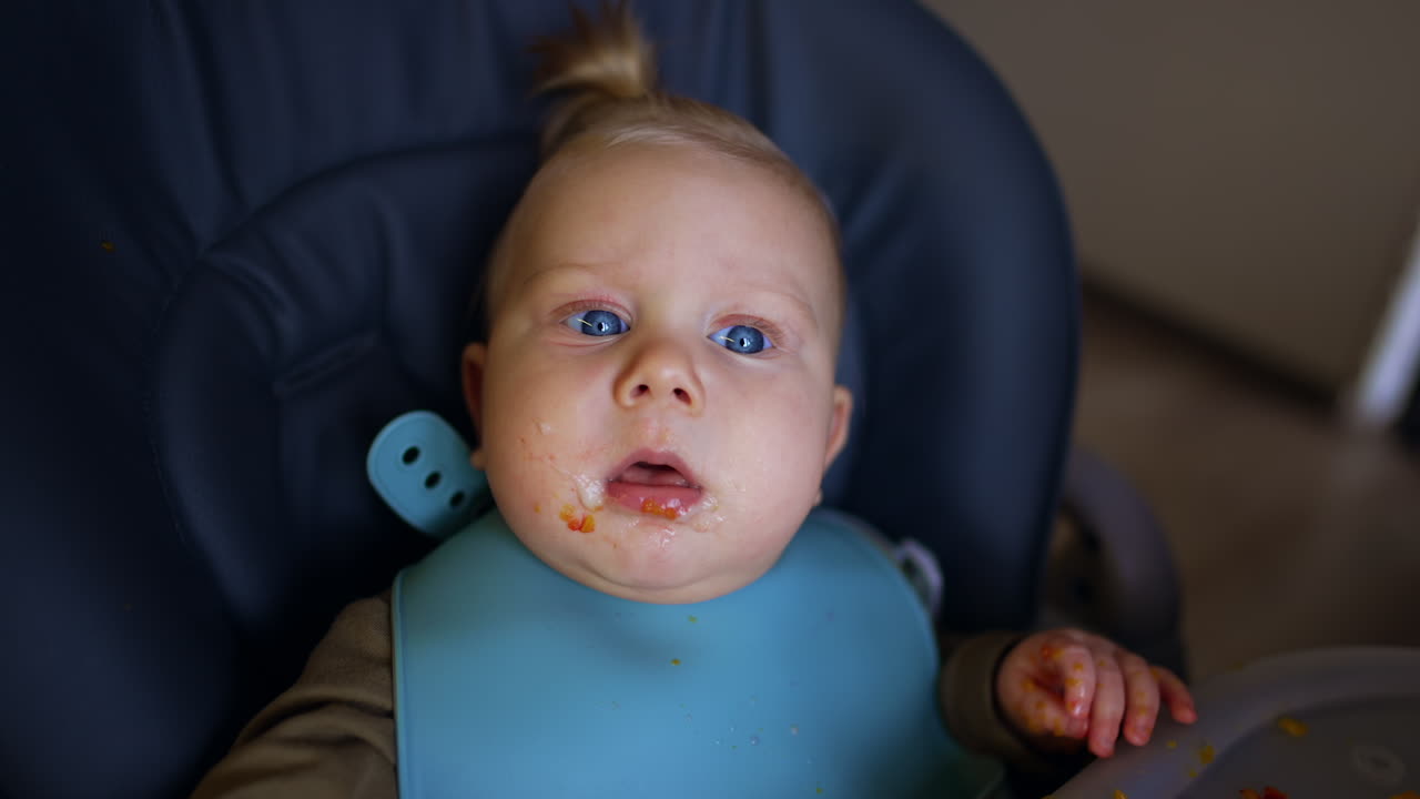 Surprised baby with food on his face looking up. Mom shoves the spoon with food to a child's mouth. Close up.