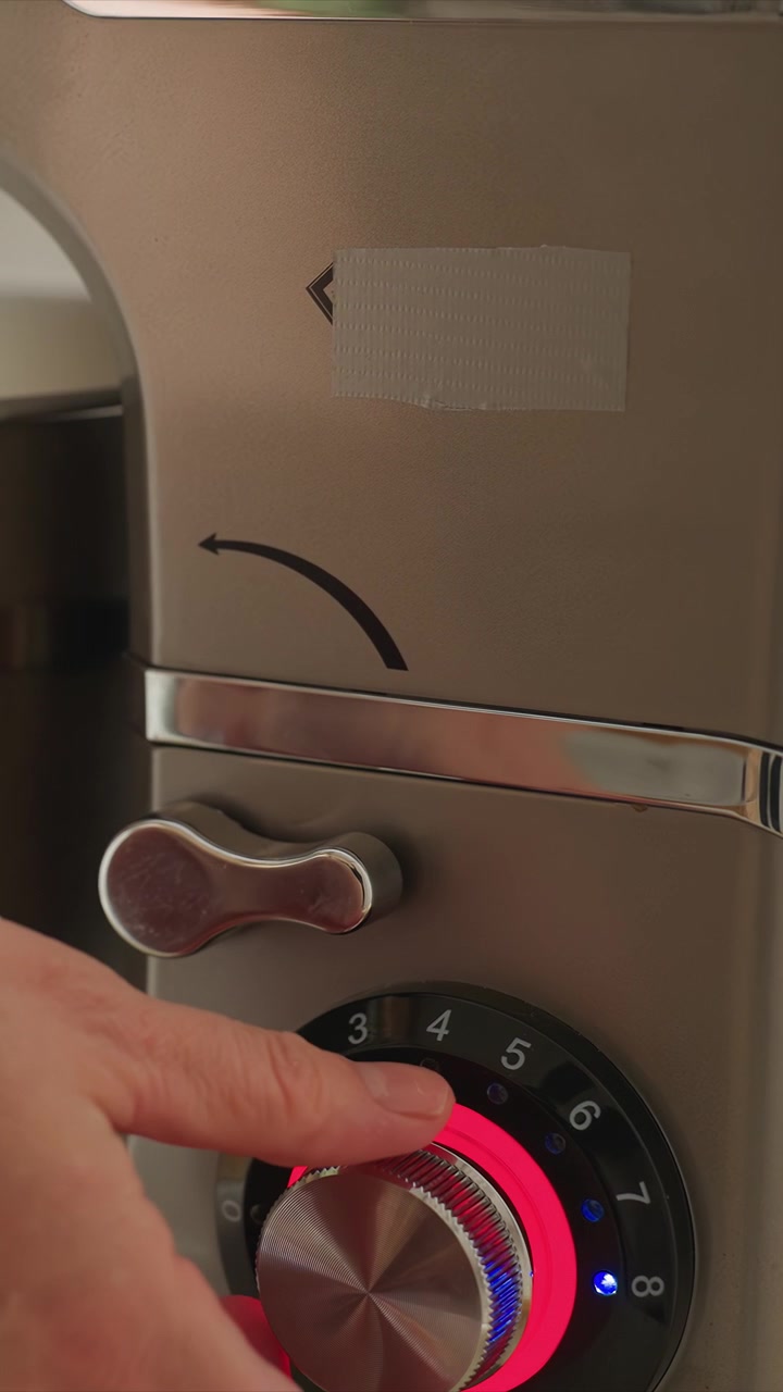 Mano con anillo de oro ajusta varios electrodomésticos de cocina. El enfoque permanece en primer plano mientras la persona interactúa con los paneles de control. Cuencos de acero inoxidable son mezclados por accesorios de batidor