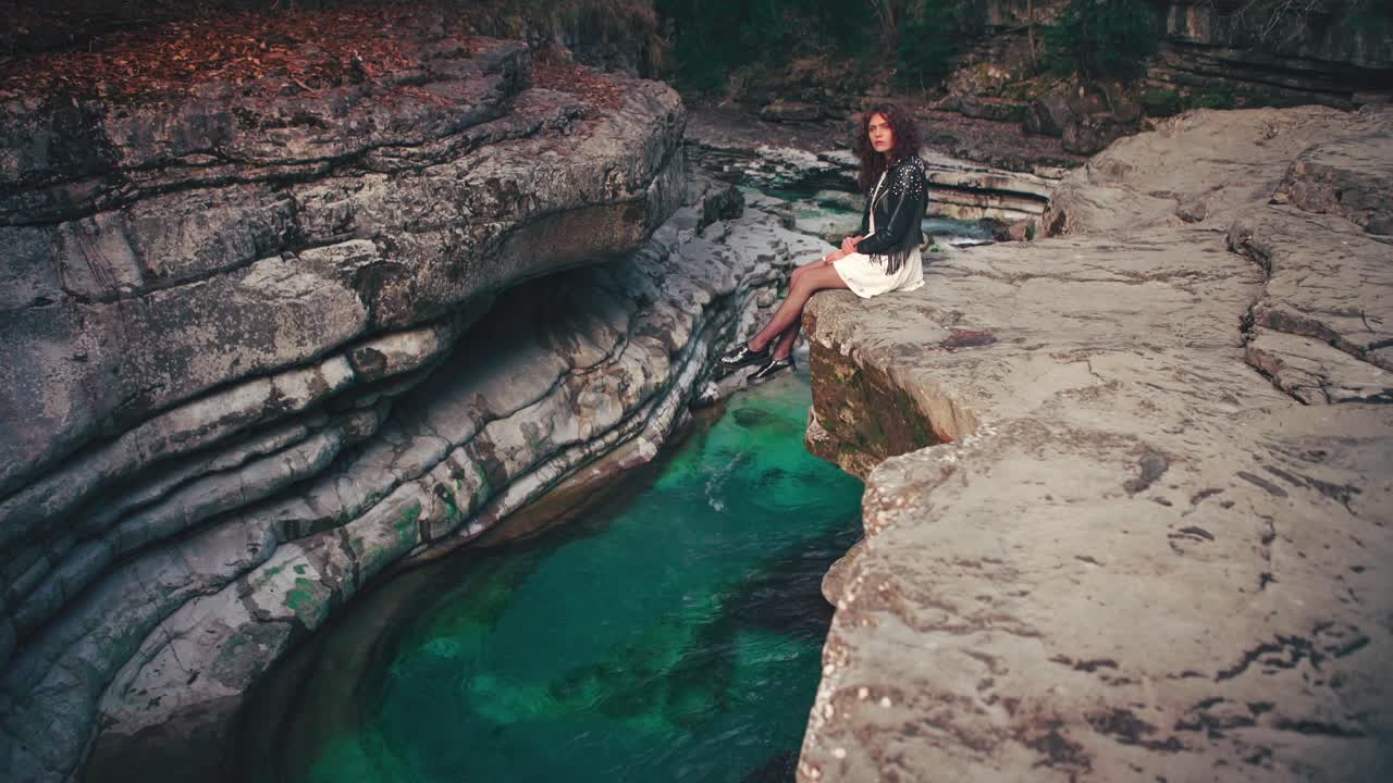 4K Cinemagraph - seamless video loop of a young brunette model woman in a white dress sitting on the edge of a gorge by a blue mountain river. The water is rushing through naturally formed rocks