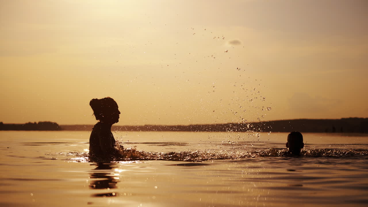 Young mother is playing with his son in water and making splash; Woman with her child is swimming in lake or river and having fun with drops; Rollick at sunset. Monochrome video. Side view.