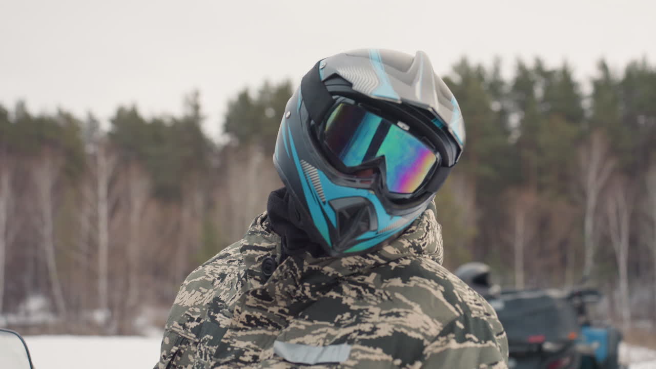Pembalap ATV menyesuaikan kaca helm, jaket kamuflase, lapangan tertutup salju, truk dan quad di latar belakang, pengendara memiringkan kepala mengamati lintasan, antisipasi yang tinggi, napas terlihat di udara dingin, siap untuk ekstrem