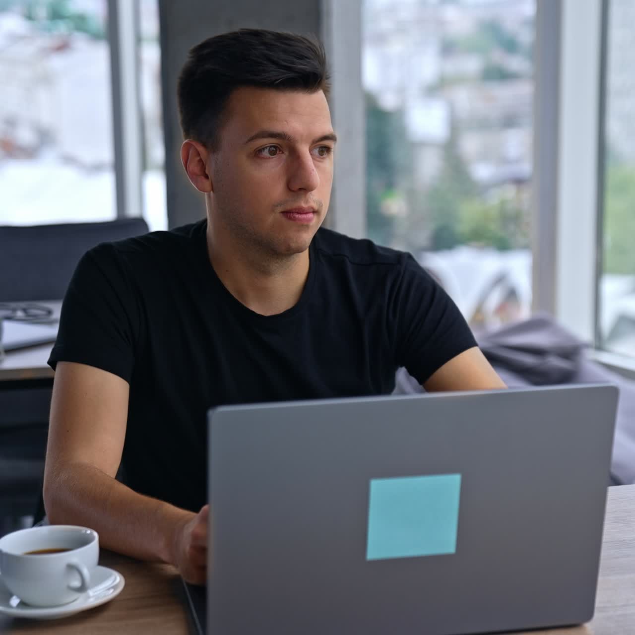Young man sitting in front of computer distracts from work. Pensive man looks away into the window. Blurred backdrop