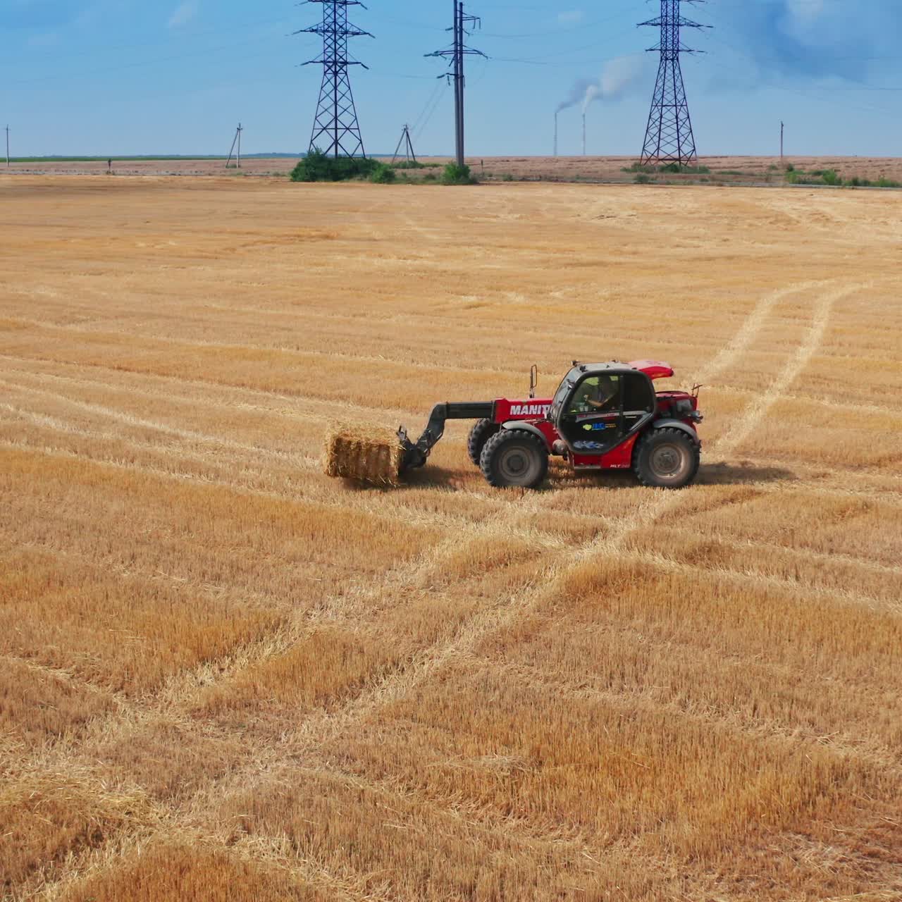 Modern red excavator lifts the bale of hay and puts it on the other bale. Electric supports and green trees at backdrop