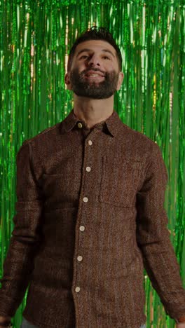 Vertical-Video-Of-Man-Celebrating-St-Patrick's-Day-Standing-In-Front-Of-Green-Tinsel-Curtain-And-Throwing-Novelty-Gold-Coins-In-The-Air