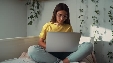 caucasian teenage girl sitting on bed and learning from laptop