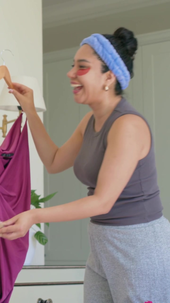 Video vertical: Mujer estirándose para elegir ropa junto a la cama, escogiendo un atuendo, sosteniendo un vestido rosa y tacones.