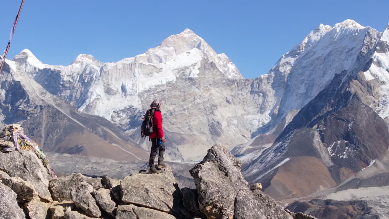 Un turista si trova sul Nangkartshang Peak nella regione dell'Everest in Nepal, bevendo acqua mentre osserva un ghiacciaio himalayano. Una ripresa da drone con effetto parallasse rivela catene montuose innevate ad alta quota.