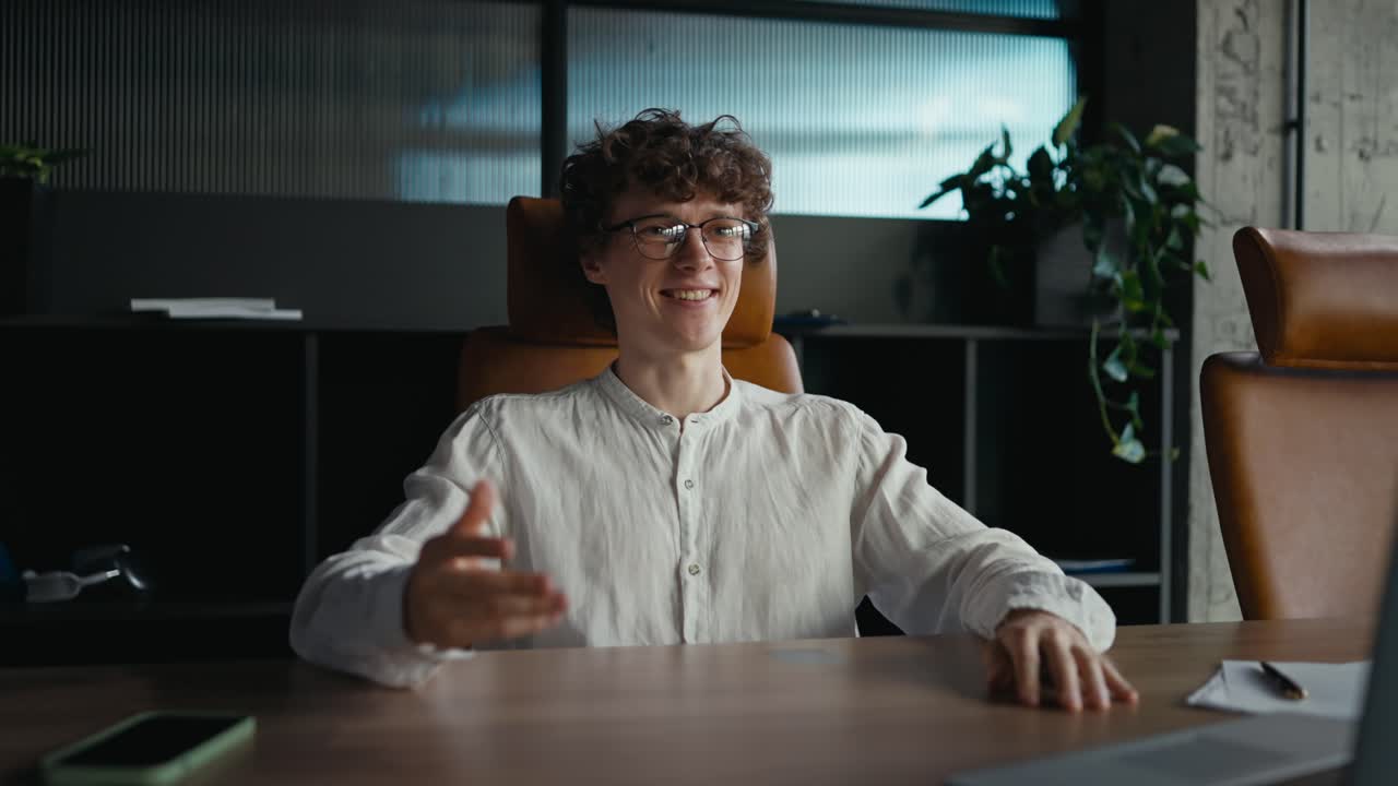 Happy guy with curly hair with glasses in a white shirt sits at the table and communicates via video conference using a laptop in the office during the working day