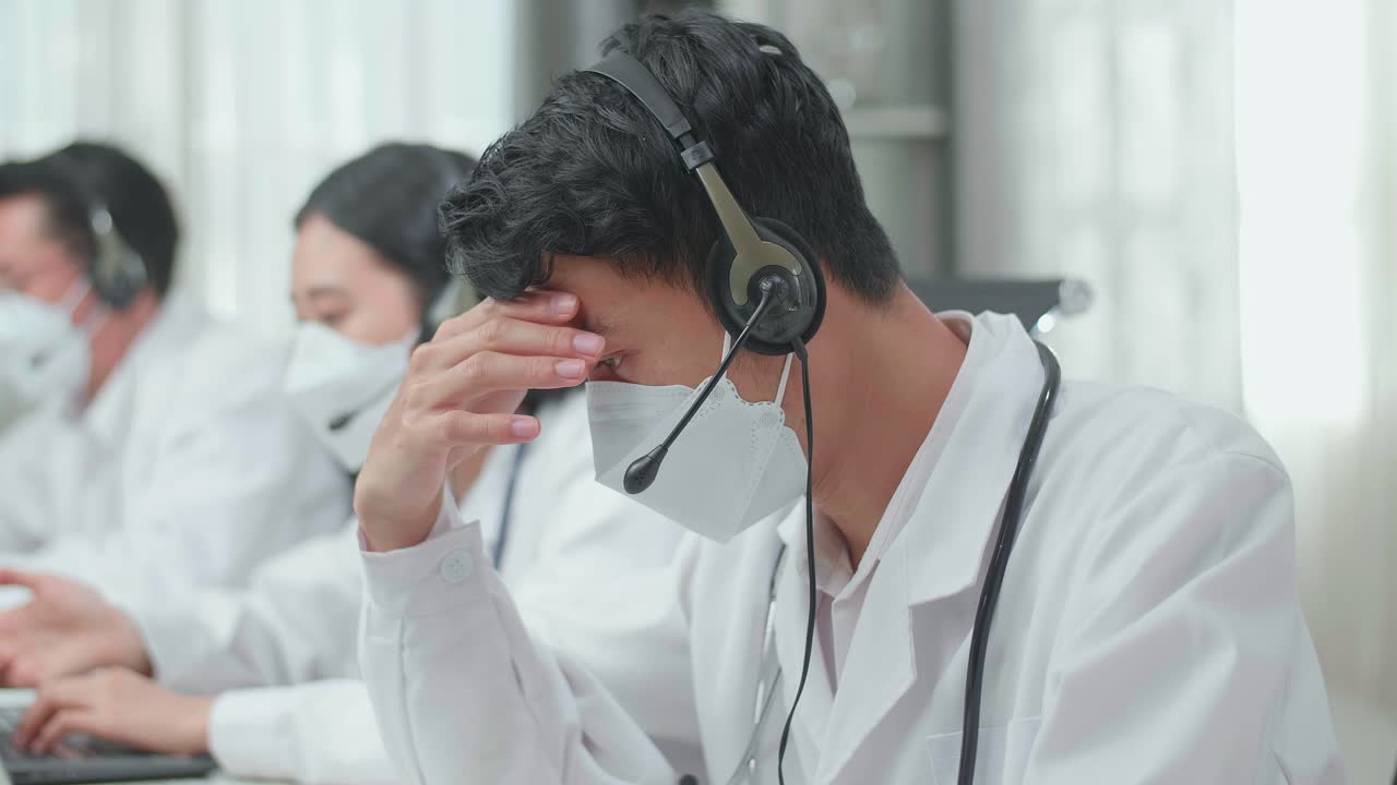 Close Up Of An Asian Man Doctor Wearing Headsets And Masks Working As Call Center Agent Is Tired Due To Working While His Colleagues Are Speaking And Typing During A Call At The Office