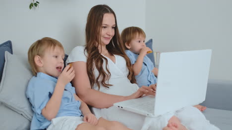 Young-beautiful-mother-and-two-young-children-sitting-on-the-couch-looking-at-the-laptop-screen-and-make-online-purchases