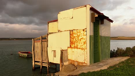 Still-shot-of-improvised-built-small-house-on-water-river-bank,-fishing-boats-floating-on-water-surface