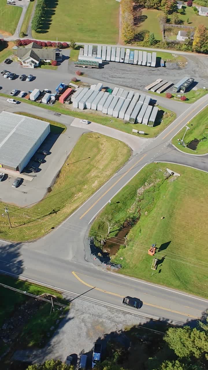 Vertikal skærm: Luftfoto af logistikplads på landet med parkerede trailere, lille lagerbygning, slynget vej, ensom sort bil, efterårstræer, inspektion af pladsansvarlig, leveringschauffør drejer