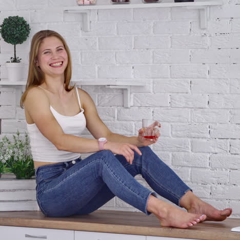 happy woman enjoys drinking fresh. side view of a girl in jeans sitting on the table in the modern kitchen. smiling female is drinking fruit juice at home.