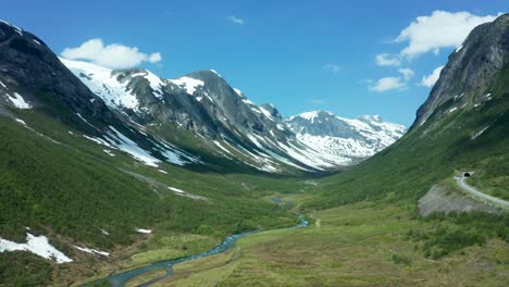 aerial pullback above lush green valleys with towering mountains in norway, under a bright sunny sky, stunning valley with cabins on grassy hill