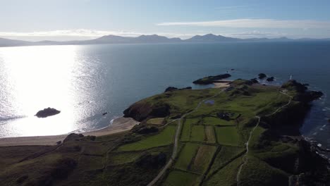 aerial pull back view ynys llanddwyn welsh island with shimmering ocean and misty snowdonia mountain range across the sunrise skyline