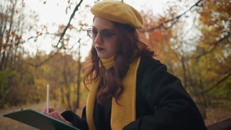 stylish lady seated focusing on her book with a green cover, adjusting her hair, surrounded by dry autumn leaves and sunlight shimmering through the trees