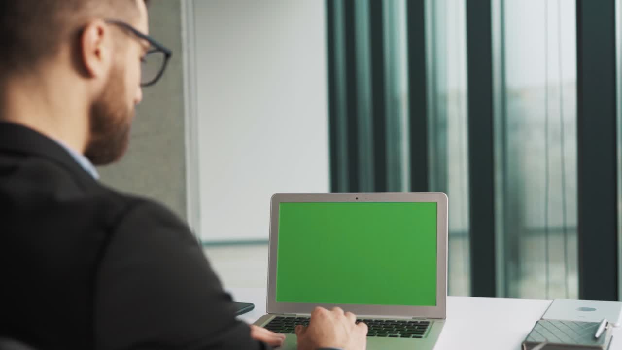 un portátil con una pantalla de cromakey sobre una mesa en una oficina con ventanas panorámicas