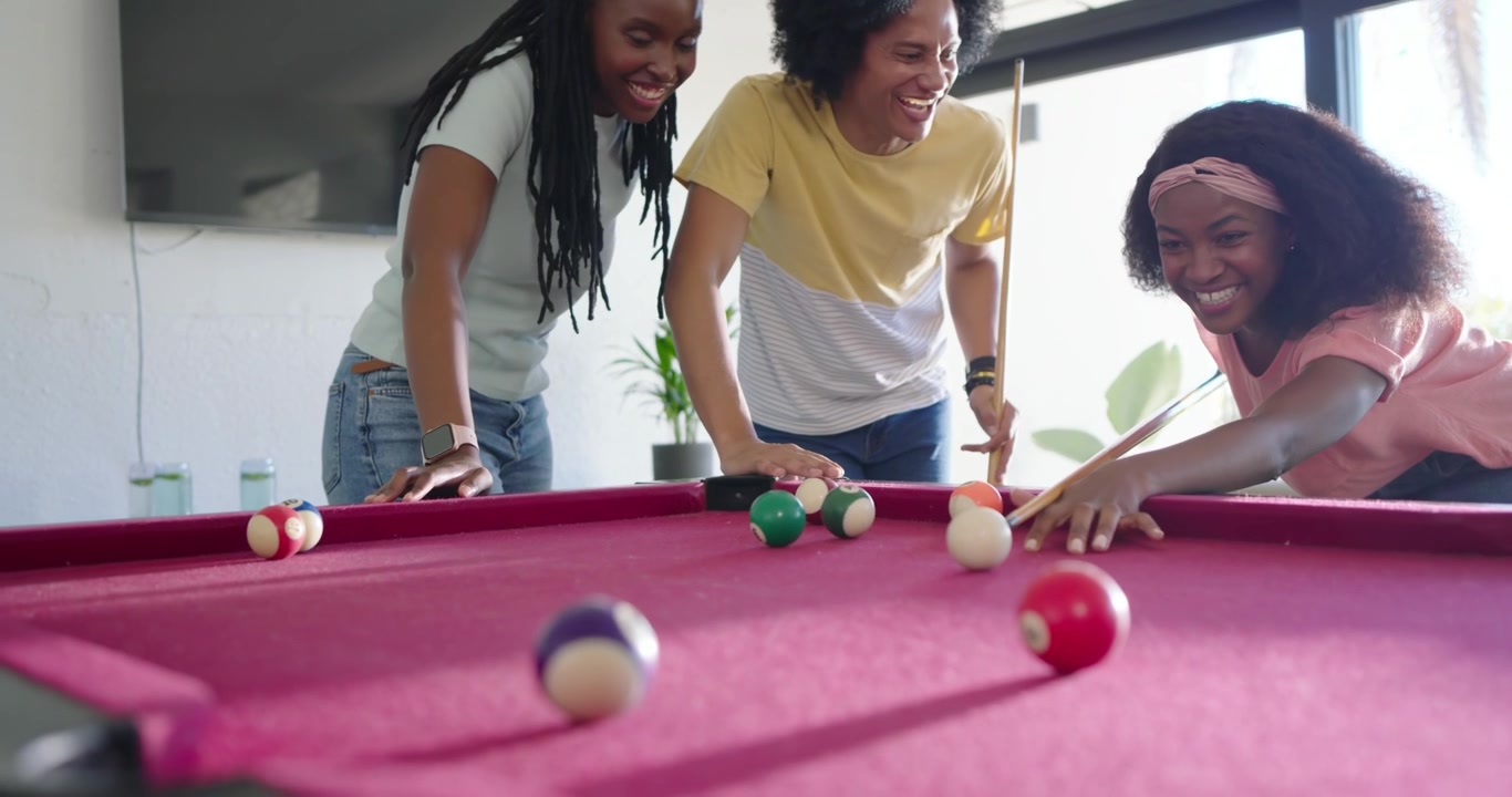 African American friends leaning at red-felt pool table, shooter striking cue ball, playing pool
