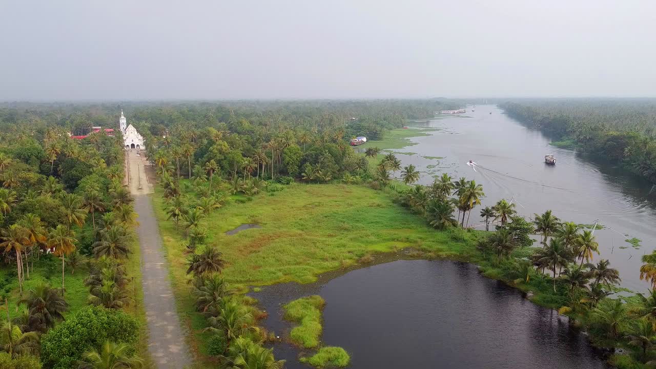 Serena bellezza di un paesaggio lacustre del Kerala. L’immagine mostra una chiesa bianca storica immersa in rigogliosi boschetti di palme da cocco, una lunga strada rurale e un fiume calmo con una barca di passaggio