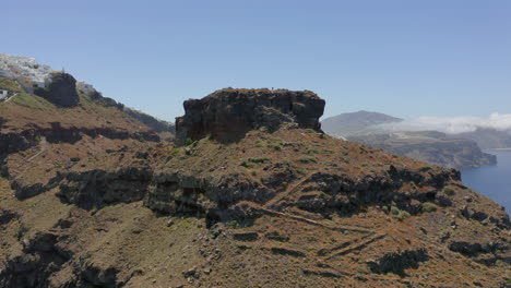 Antena:-Sobrevuelo-Panorámico-De-Un-Hombre-Caminando-Sobre-La-Roca-Skaros-En-Santorini,-Grecia