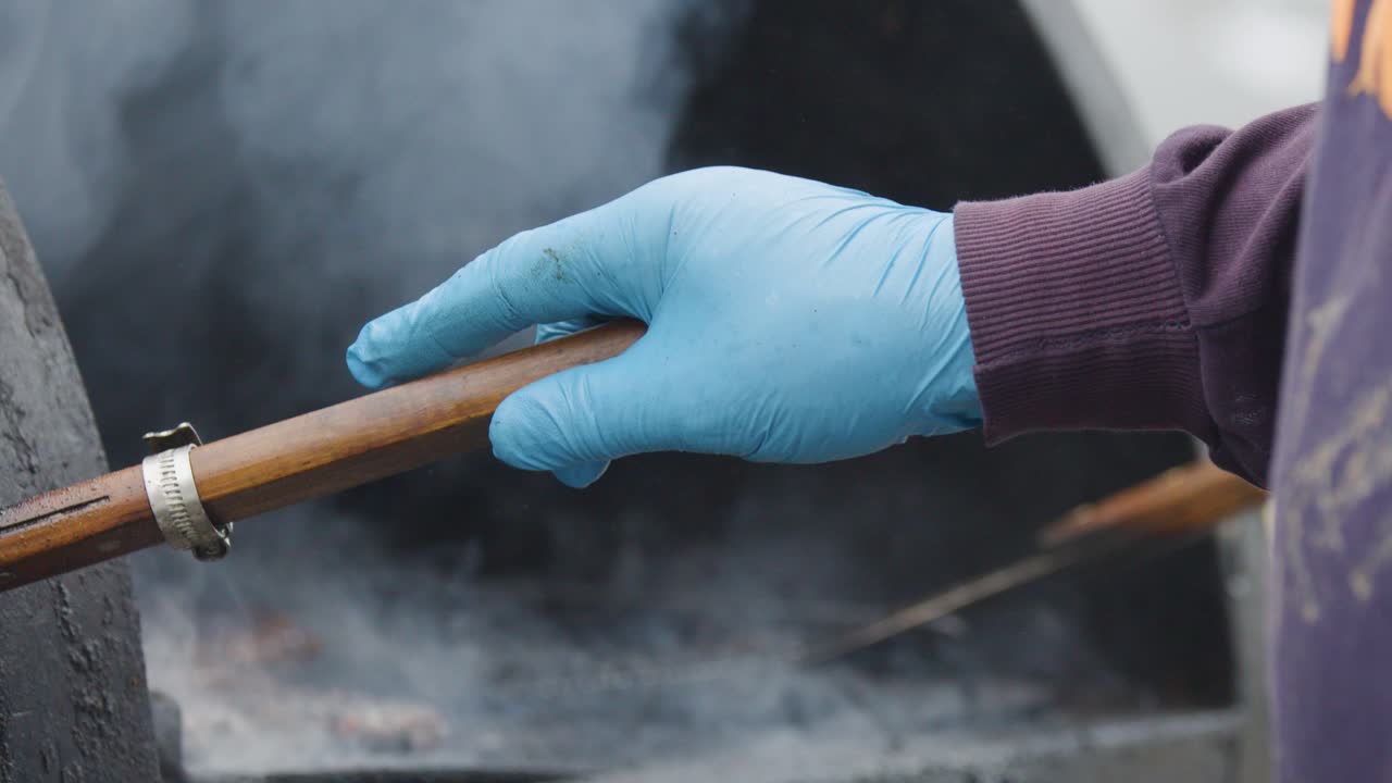 A person wearing blue gloves adjusts a wooden-handled vent on a large outdoor barbecue smoker, surrounded by thick smoke in natural daylight with close-up camera angles