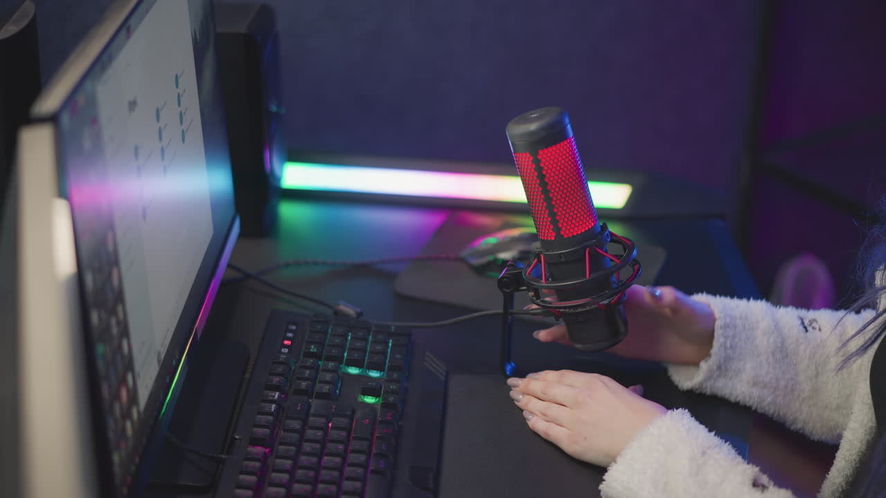 tech lady in fluffy sweater reaches to adjust desktop condenser mic position and cable next to rgb lit monitor keyboard preparing livestream recording setup in home office with pop filter backdrop