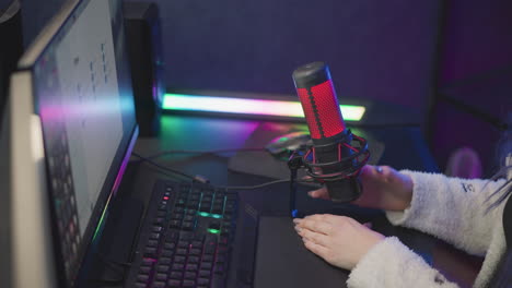 tech lady in fluffy sweater reaches to adjust desktop condenser mic position and cable next to rgb lit monitor keyboard preparing livestream recording setup in home office with pop filter backdrop