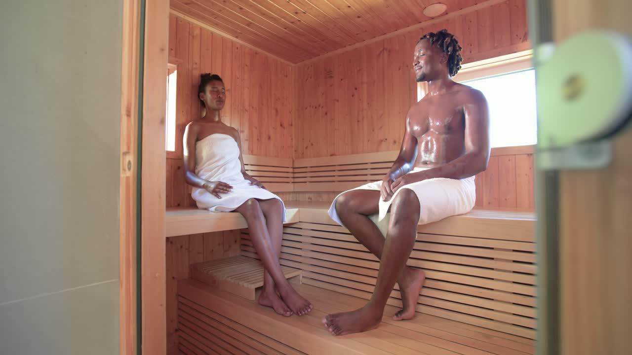 Coppia afroamericana che si rilassa su panche da sauna in asciugamani, la donna che guarda, l'uomo che si gira ridendo