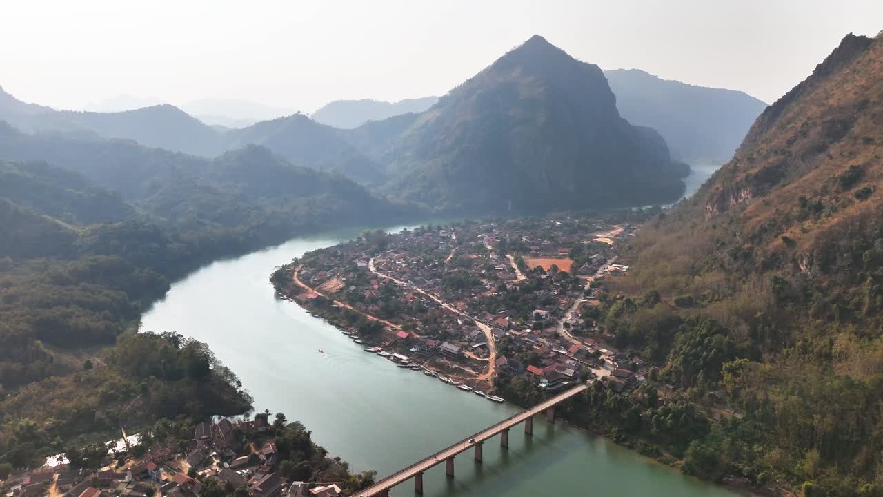 Drone view over Nong Khiaw town along the Nam Ou River in northern Laos, with the concrete Nong Khiaw Bridge visible and karst peaks of Phou Pha Daeng rising in the distance under hazy sky