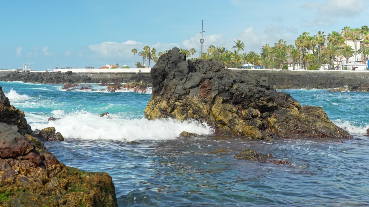 Rocce e mare con foresta di palme e cittadina sullo sfondo, al rallentatore.